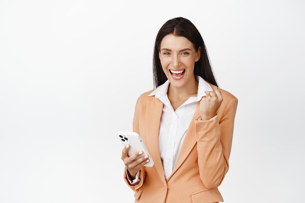 スマートフォンを持って、ガッツポーズを作って笑顔で成功したセールスウーマンは、白い背景の上に立っている携帯電話で勝利を喜んでいます