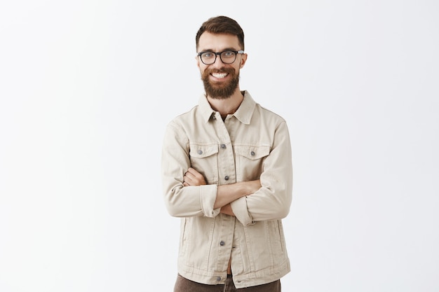 Successful smiling bearded man in glasses posing against the white wall