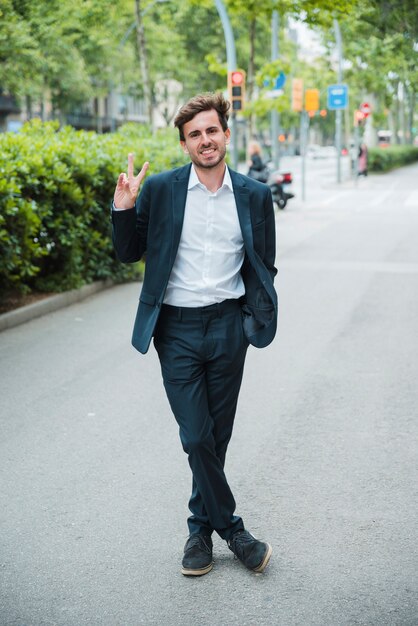 Successful smiling young businessman standing on street showing victory sign