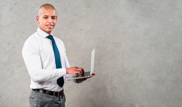 Free photo successful smiling young man using laptop looking to camera standing against grey wall