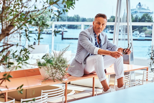 Foto gratuita uomo alla moda di successo vestito con abiti eleganti moderni seduto su un tavolo al caffè all'aperto sullo sfondo del molo della città.