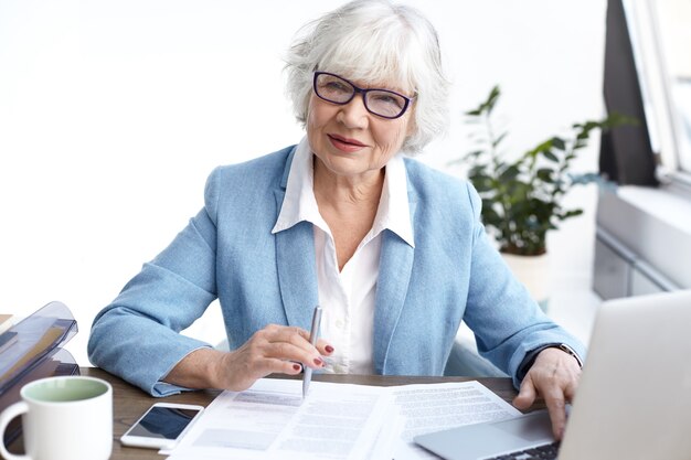 Successful stylish mature female Chief Executive Officer wearing eyeglasses and formal clothes looking through financial report, working at office desk, using electronic gadgets and making notes