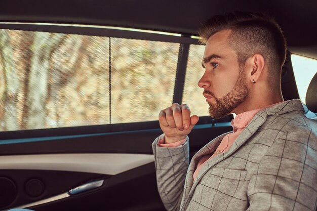 Успешный стильный молодой бизнесмен в сером костюме и пинг-рубашке едет на заднем сиденье роскошного автомобиля.