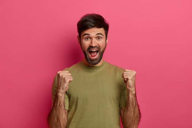 Successful teen boy raises clanched fists, celebrates triumph, looks with joy, exclaims loudly, has thick stubble, wears casual t shirt, poses over pink wall screams yes, got prize, wins contest