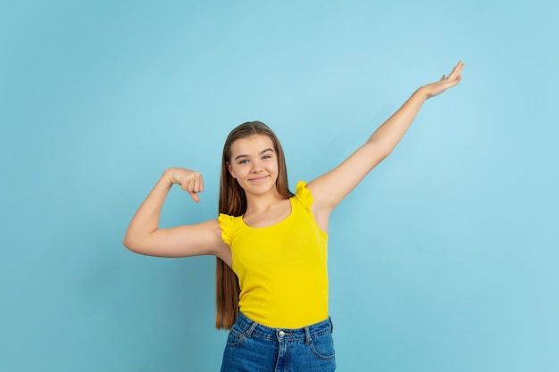 Successful winner. Caucasian teen girl's portrait isolated on blue wall. Beautiful model in casual yellow wear. Concept of human emotions, facial expression, sales, ad. Copyspace. Looks cute.