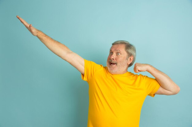 Successful winner gesture. Caucasian man portrait isolated on blue studio background. Beautiful male model in yellow shirt posing. 