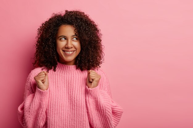 Successful young African American teenage girl celebrates accomplishment, raises clenched fists, dressed in oversized winter sweater, smiles broadly, looks aside, isolated over pink background.