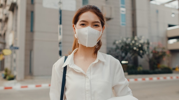 Успешная молодая азиатская коммерсантка в модной офисной одежде в медицинской маске, улыбаясь на улице