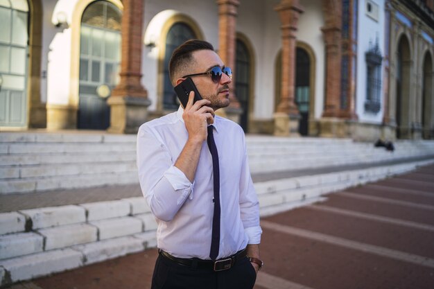 Успешный молодой бизнесмен в официальном наряде с солнцезащитными очками разговаривает по телефону