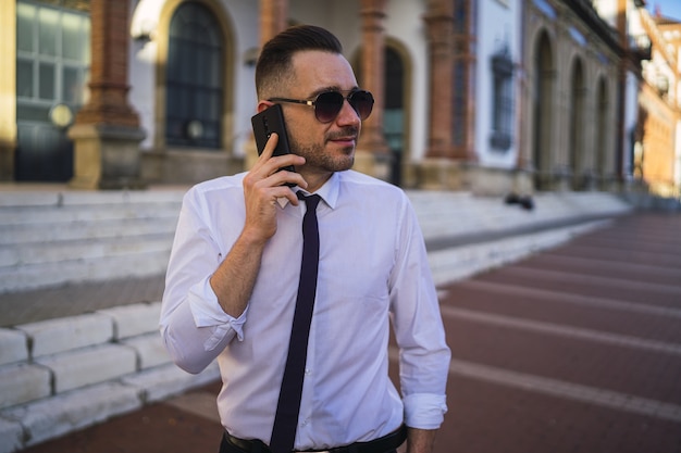Успешный молодой бизнесмен в официальном наряде с солнцезащитными очками разговаривает по телефону