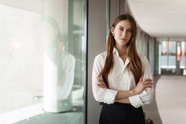 Successful young businesswoman, standing near the window in office corridor, smiling and looking camera.