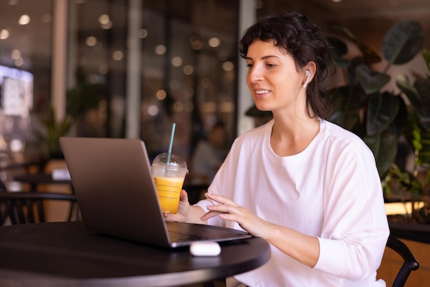Successful young caucasian brunette woman in white tshirt uses headphones and laptop sitting at table in cafe