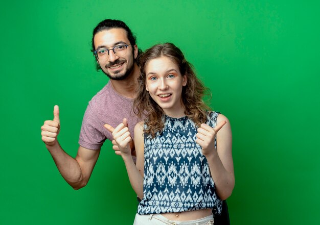 Successful young couple man and woman looking at camera smiling cheerfully showing thumbs up over green background