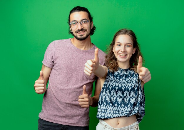 成功した若いカップルの男性と女性は元気に緑の壁に親指を見せて笑っている
