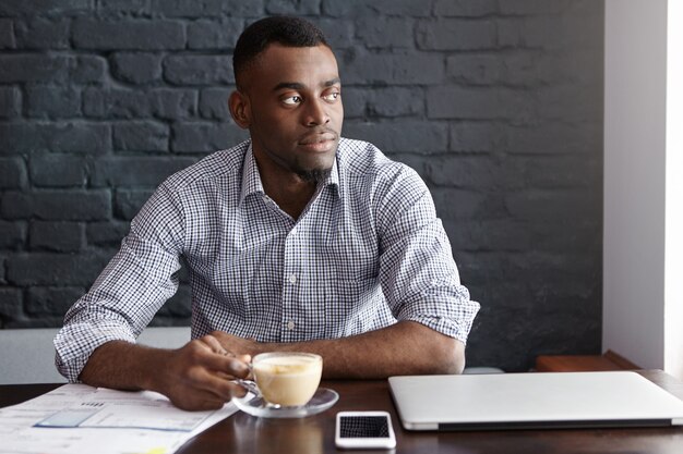 Successful young dark-skinned CEO having his morning coffee, sitting at cafe table with laptop