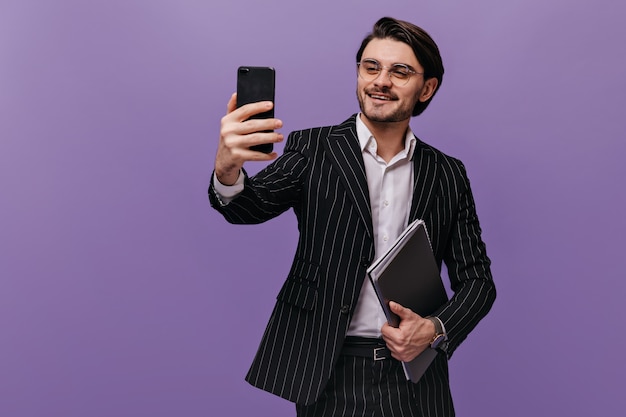 Successful young gentleman in white shirt, black striped suit and trendy glasses making selfie with folders and smiling