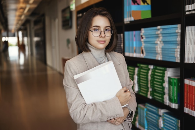 Free photo successful young woman enrolled in university, studying hard get degree, working on project, holding documents and smiling camera with relaxed expression, standing near piles of books in hall.