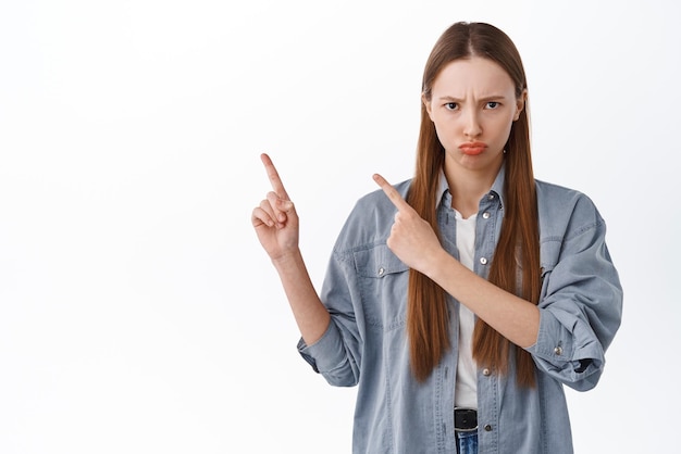 Sulking teenage girl pointing aside with jealous unfair face complaining looking displeased and frowning pouting at camera standing against white background