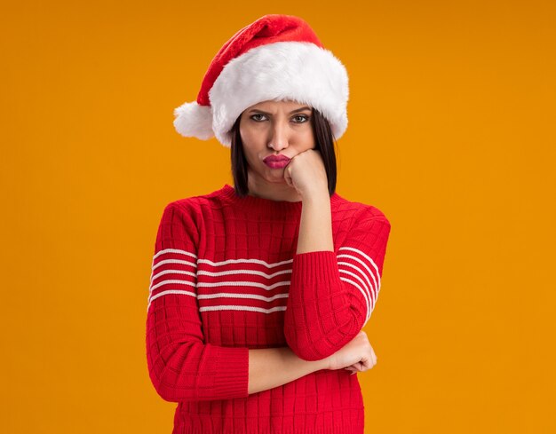 Sulking young girl wearing santa hat looking at camera keeping hand on chin isolated on orange background with copy space