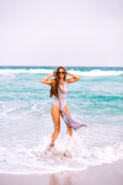 Summer beach portrait of pretty brunette woman with sportive perfect tanned body and long brunette hairs, wearing trendy glamour elegant swimsuit, model relax near ocean, spin and having fun, freedom.