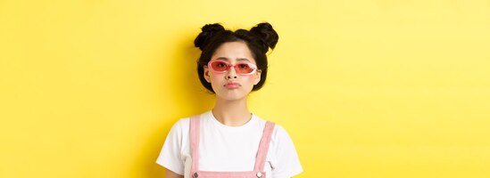 Free photo summer and fashion concept bored asian teen girl in sunglasses pouting standing moody on yellow back