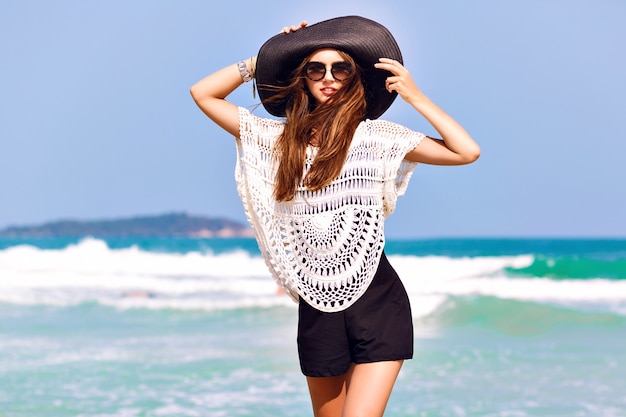 Summer fashion portrait of beautiful woman enjoy windy sunny day near ocean, vacation style. Young stylish girl wearing black romper vintage hat and big sunglasses, bright colors