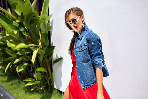 Summer fashion portrait o stunning elegant fashion girl posing outside in tropical country, wearing elegant luxury dress and trendy denim jacket, dancing and having fun.