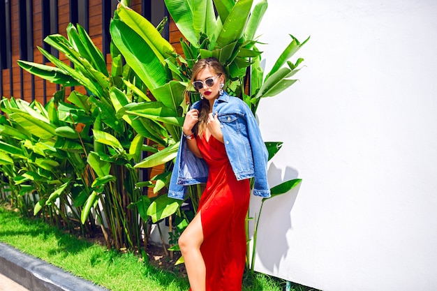 Summer fashion portrait of pretty blonde girl having fun on tropical vacation posing near palm leaves, wearing maxi red silk dress and trendy hipster denim jacket, dancing alone.