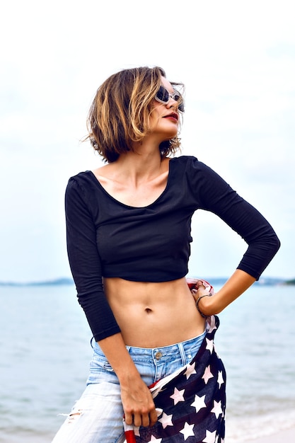 Летняя мода портрет потрясающе подтянутой сексуальной женщины в джинсовой ткани и укороченном топе, держащей американский флаг, проводит время на пляже в дождливый день.