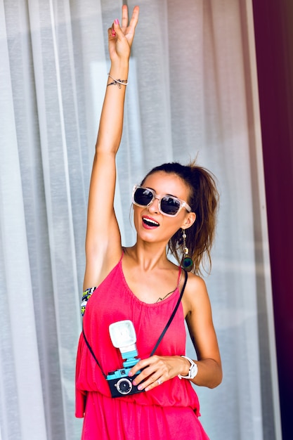 Summer fashion portrait of young woman posing smiling and having fun, wearing neon stylish jumpsuit and sunglasses, holding funny retro camera. showing piece science.