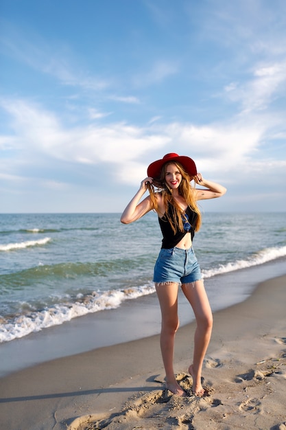 바다 근처에서 혼자 걷는 젊은 여성의 여름 패션 초상화, 해변에서의 휴가, 혼자 여행, 빈티지 모자 선글라스와 데님 반바지, 슬림 바디, 일출, 건강한 라이프 스타일을 입고.
