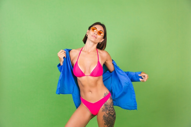 Summer fit sporty woman in pink bikini, blue shirt and orange sunglasses on green, happy cheerful joyful positive