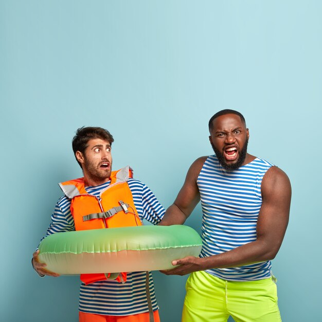 夏の休日のコンセプト。 2人の男性のショットは、膨らんだ浮き輪を共有できません。怒っている暗い肌の男は、ビーチのライフガードに水泳用具を要求します