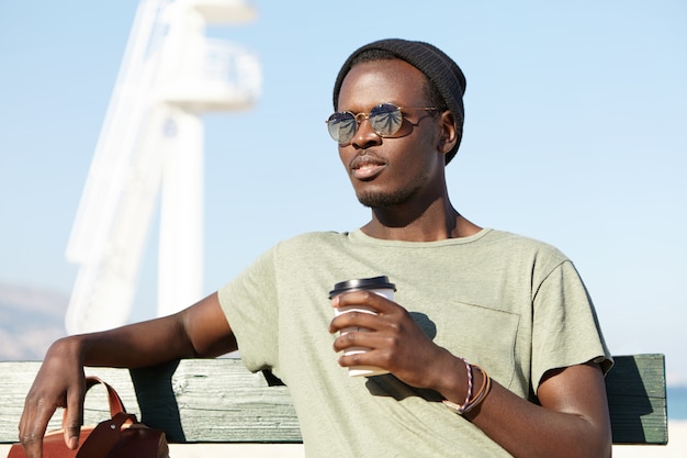 Summer, holiday and vacation. Outdoor shot of relaxed carefree dark-skinned male tourist in eyewear and hat sitting on wooden bench