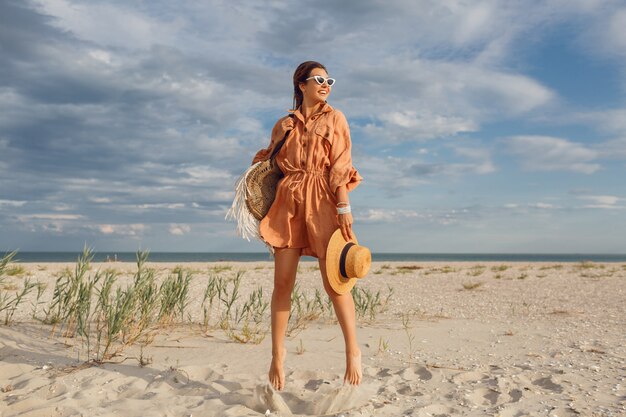 Summer image of beautiful brunette female in trendy linen dress jumping and fooling around , holding straw bag. Pretty slim girl enjoying weekends near ocean. Full length.
