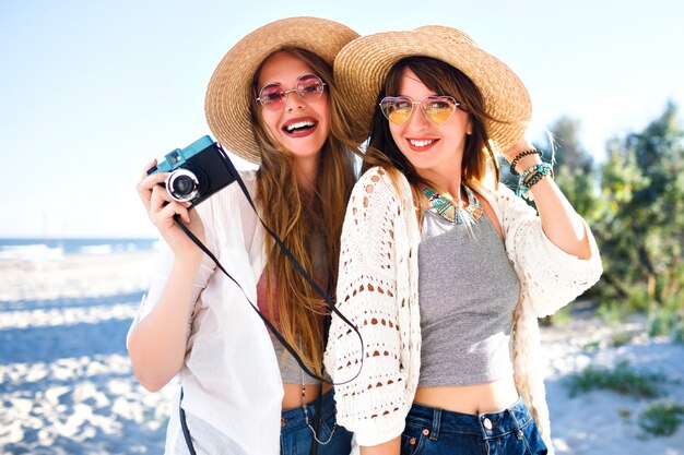 Summer lifestyle portrait of happy best friends sister girls posing on the beach, sunny light colors,  straw hats and sunglasses, holding vintage photo camera, having fun together.