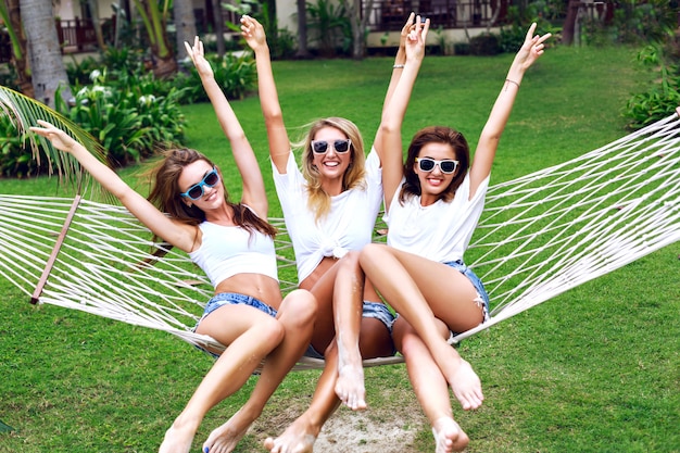 Summer lifestyle portrait of tree women going crazy, screaming, laughing having fun together, jumping at hammock. wearing white tops and sunglasses, ready for party, joy, fun.