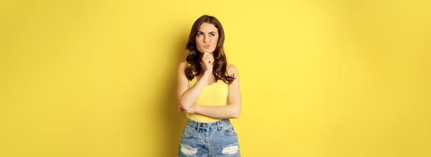 Free photo summer and lifestyle thinking girl looking aside and deciding making choice standing thoughtful wearing tank top posing against yellow background