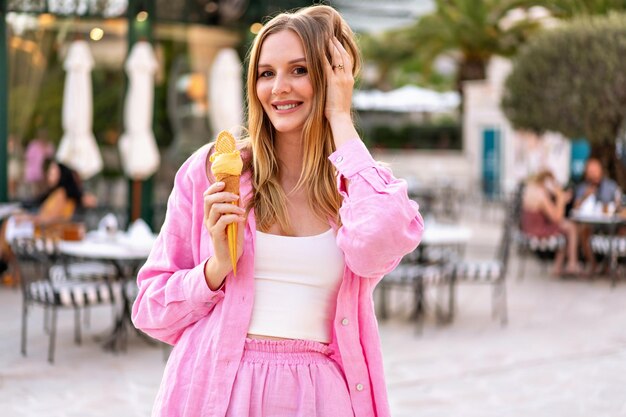 Summer outdoor portrait of pretty blonde woman eating tasty Italian gelato cone ice cream enjoy her vacation pink trendy outfit