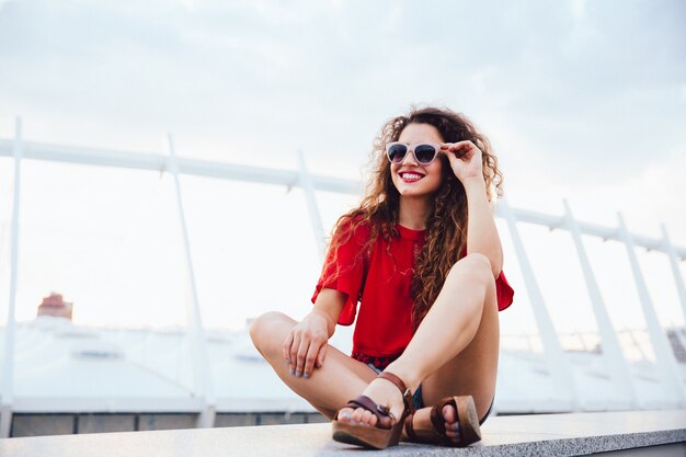 Summer photo of attractive funny girl in sunglasses with curly hair sitting alone