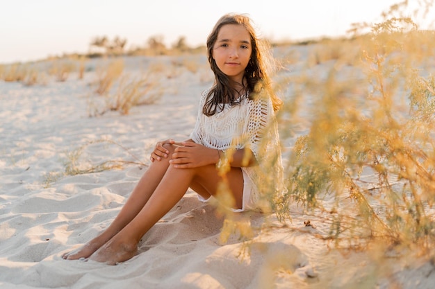 Summer photo of small girl in stylish boho outfit posing on the beach Warm sunset colors Wacation and travel concept