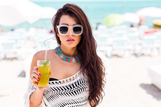 Summer portrait of beautiful brunette woman chilling in the beach club. Tropical acsessories.