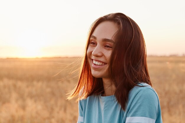 Летний портрет смеющейся счастливой женщины на открытом воздухе, наслаждающейся теплым солнцем, в синей футболке, с темными волосами, глядя в сторону с зубастой улыбкой, выражающей счастье.
