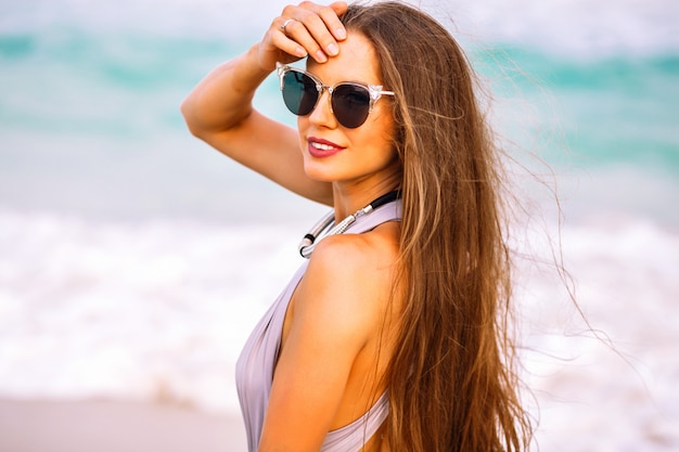 Summer portrait of sensual pretty woman posing near ocean, long hairs, luxury trendy bikini and accessories, vacation time.