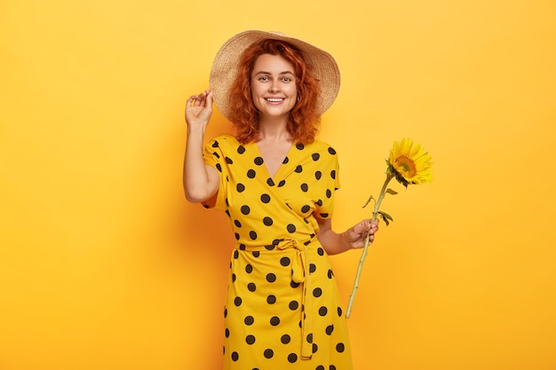 Summer portrait of smiling mirthful ginger lady with tender smile, stands with sunflower picked from field