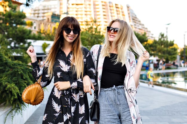 Summer portrait of two stylish pretty best friends woman having great time together, smiling and enjoy time on the street, elegant trendy clothes begs and sunglasses, happy couple, relationships .