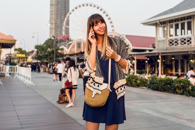 Summer  positive portrait of cheerful woman in stylish  outfit talking by mobile phone  and smiling on  Riverfront in Bangkok