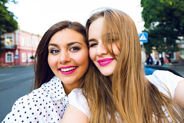 Summer sunny image of two sisters best friends brunette and blonde girls having fun on the street, making selfie, , bright stylish make up long hairs
