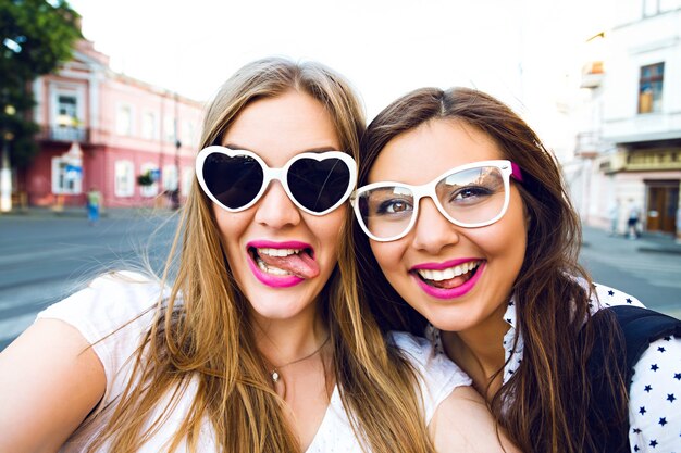 Летний солнечный образ двух лучших подруг сестер, брюнетки и блондинки, веселых на улице, делающих селфи, в забавных винтажных солнцезащитных очках, яркого стильного макияжа с длинными волосами
