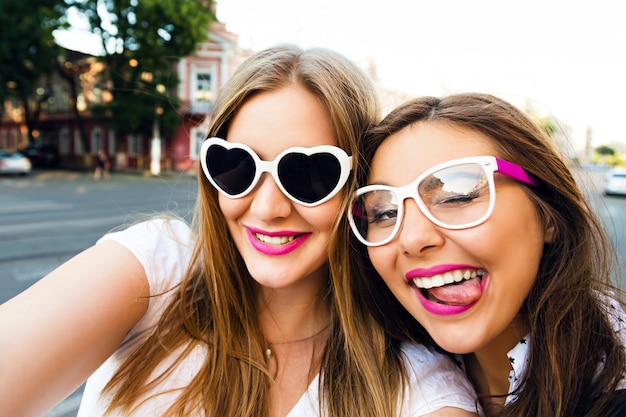 Summer sunny image of two sisters best friends brunette and blonde girls having fun on the street, making selfie,wearing funny vintage sunglasses, bright stylish make up long hairs